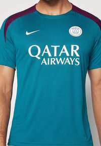 Teal Nike-paita, jossa on viininpunaiset olka-akselit. Rintamuksessa on valkoisella painettu "QATAR AIRWAYS" ja Paris Saint-Germain -logo.