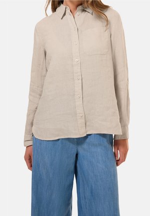 Woman wearing a beige long-sleeve button-up shirt with a chest pocket and loose blue pants against a plain background.