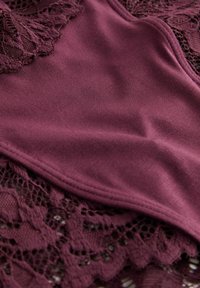Close-up of burgundy fabric with smooth, solid section overlaying intricate floral lace pattern.