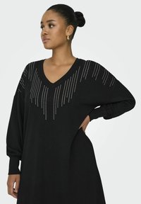 Abito nero oversized con scollatura a V, decorato con linee verticali di borchie argentate nella zona del petto. Maniche lunghe e gonfie.