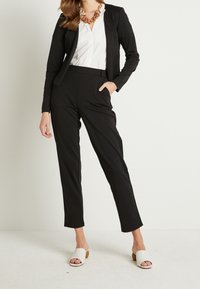 Blazer nero e pantaloni neri abbinati realizzati in tessuto liscio, abbinati a una camicia bianca e mule aperti di colore chiaro.