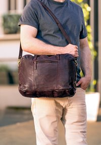 Brown leather messenger bag with zippered pocket, dual handles, and adjustable shoulder strap. Features smooth texture and rounded edges.