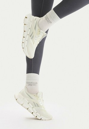 Person trägt weiße Reebok-Sneaker, weiße Reebok-Socken und schwarze Leggings, wobei ein Bein gegen einen weißen Hintergrund angewinkelt ist.