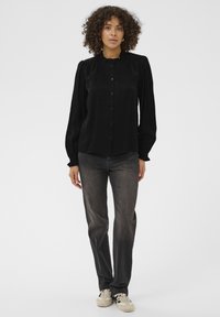 Musta pitkähihainen paita, jossa on pyöreä kaulus ja nappikiinnitys, yhdistettynä tummanharmaisiin farkkuihin ja vaaleanbeigeihin tennareihin. Silkkinen kangas, jossa on hieno kiilto.