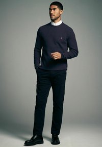 Mørkeblå sweater i et struktureret strikdesign med ribbede manchetter, parret med mørke bukser og sorte sko. Bemærkelsesværdigt rødt logo på brystet.