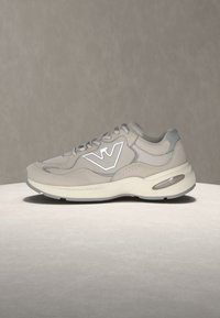 Baskets gris clair et beige avec semelle blanche, logo de marque sur le côté, dessus en mesh et cuir, posées sur une surface neutre avec un fond uni.