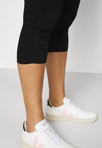 Mustat capri-housut, joissa on röyhelöillä varustetut sivusaumat, yhdistettynä valkoisiin tennoihin, joissa on vaaleanpunainen logo ja musta kantapääkorostus.