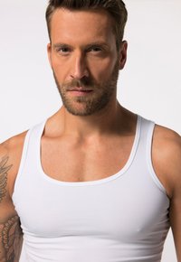 Hvid bomulds tanktop på en mand, med en rund halsudskæring og brede skulderstropper. Nærbillede af overkroppen og ansigtstræk.