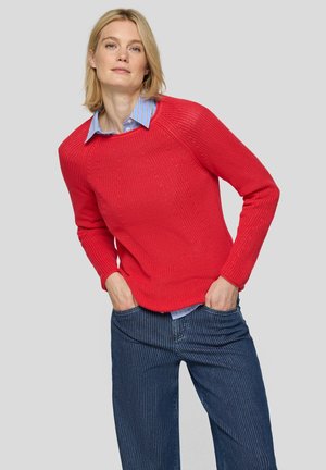 Woman with blonde hair wearing a bright red knit sweater over a blue striped button-up shirt and blue pinstriped pants with hands in pockets.