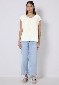 Top giallo a maniche corte con scollatura a V, abbinato a jeans azzurri a gamba larga e sneakers grigie con accenti neri.