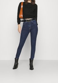 Jeans skinny de talle alto de color azul oscuro, suéter negro corto, botines negros con tacón hasta el tobillo y un bolso de hombro de piel de cocodrilo marrón con una correa larga.