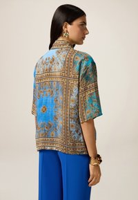 Bluză ușoară albastră cu modele florale intricate maronii, mâneci scurte și croială lejeră, asociată cu pantaloni albaștri.