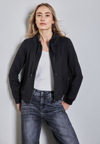 Veste noire zippée avec un col montant, des poignets côtelés et des poches, portée sur un haut blanc et associée à un jean en denim foncé.