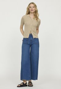 Cardigan en tricot beige à manches courtes, associé à un jean en denim bleu à jambes larges avec poches avant et ourlet effiloché. Des sandales noires viennent compléter le look.