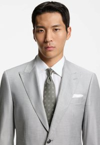 Lichtgrijs colbert met gereviseerde revers, gecombineerd met een wit overhemd en een donkergroene stropdas met patroon. Pochet zichtbaar in het colbert.