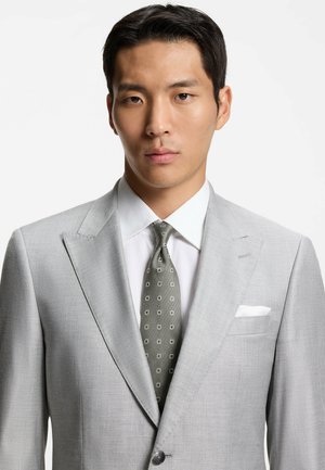 Veste de costume gris clair à revers crantés, associée à une chemise blanche et une cravate vert foncé à motif. Pochette visible dans la veste.