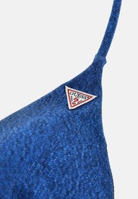 Blue denim bralette with a smooth texture, featuring a triangular red and white "GUESS" logo patch on the left. Thin shoulder straps.