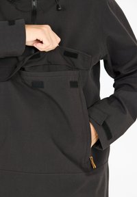 Veste noire avec une poche avant à fermeture Velcro, accent zipper et manches ajustables. Tissu durable avec une texture mate.