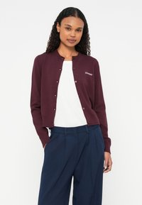 Cardigan bordeaux con colletto rotondo e chiusura a bottoni, stoffa morbida, polsini a coste, con logo "2ndDay" sul lato sinistro del petto. Abbinato a pantaloni blu navy.