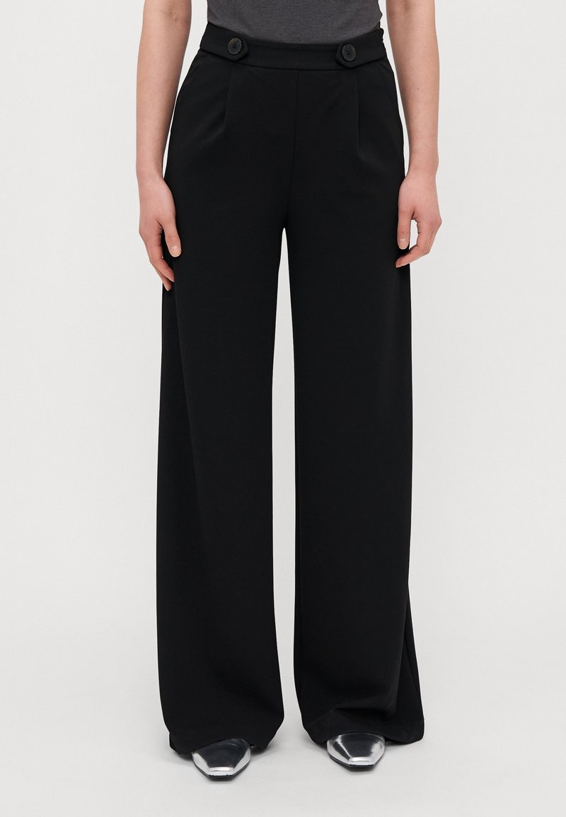 Pantalon noir taille haute à jambes larges avec deux grands boutons à la taille, porté avec des chaussures pointues argentées et un haut gris.