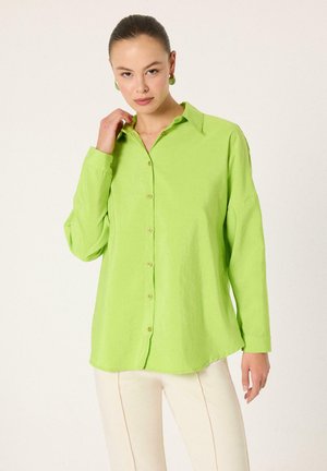 Nő élénkzöld gombos ingben és bézs nadrágban, tiszta világos háttér előtt áll, és az egyik kezével a gallérját érinti.