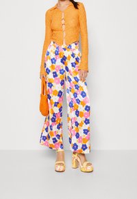 Top de encaje naranja brillante con detalles de botones, pantalones de pierna ancha con estampado floral en rosa, azul y blanco, y sandalias de plataforma amarillas.