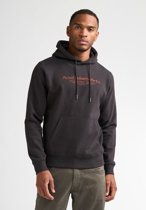 Man die een zwart hoodie met oranje tekst en groene broek draagt, staand tegen een effen lichte achtergrond.