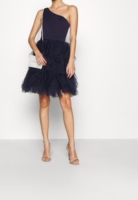 Yhden olkapään tummansininen mekko, jossa on vartalonmyötäinen yläosa ja kerroksittain laskostettu tyllihame. Yhdistetty pieneen helmikoriseen käsilaukkuun ja beigeihin korkokenkiin.