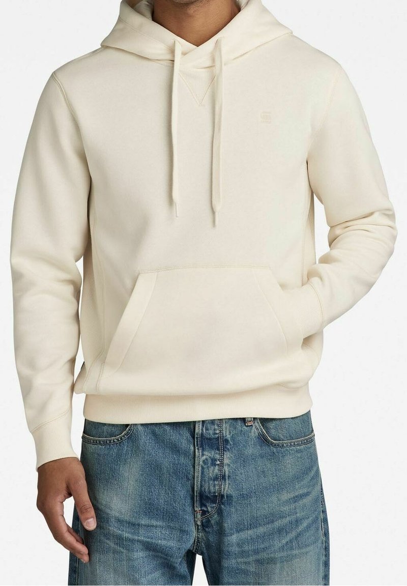 Homme portant un sweat à capuche couleur crème avec poche avant et cordons, associé à un jean denim bleu, debout avec une main dans la poche.