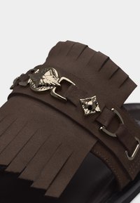 Bride en daim marron avec détails à franges et ornements en métal doré en forme d'oiseau et de boucle sur semelle noire.