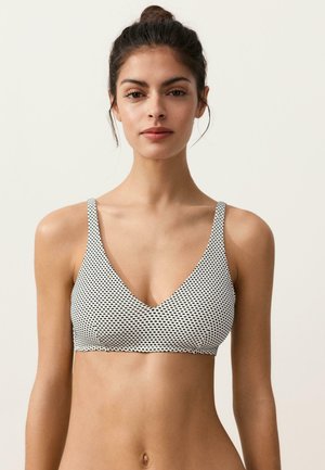 Młoda kobieta o ciemnych włosach związanych w kok, ubrana w białą i czarną wzorzystą bezrękawnikową braletkę, stojąca na tle jednolitego jasnego tła.