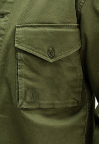 Olijfgroene shirt met een knoopzak op de borst met een klep, textuurstof en een genaaid logo-detail. Gladde oppervlakte en gestructureerd ontwerp.