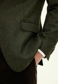 Blazer im Fischgrätenmuster aus dunkelgrünem Wolle, mit Reverskragen, einer einzelnen Brusttasche und einem weißen Hemdmanschette, die hervorblitzt.