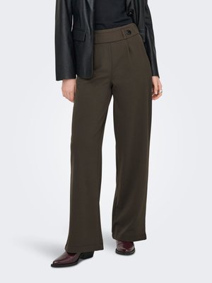 Persona que lleva pantalones anchos de color oliva oscuro, chaqueta de cuero negra, camiseta negra y botas burdeos con punta afilada, de pie sobre fondo blanco.
