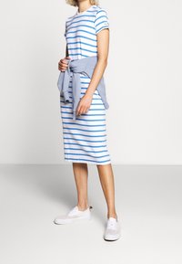 Donna che indossa un abito midi a righe bianche e blu con una felpa azzurra chiara annodata in vita e sneaker bianche.