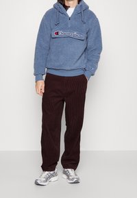 Modrá fleecová mikina s půlzipem a vyšívaným logem, v kombinaci s hnědými manšestrovými kalhotami a stříbrnými sportovními botami. Neformální outfit.