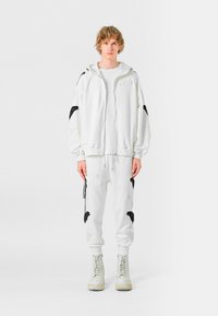 Sudadera blanca con capucha y bordado de alas negras, combinada con pantalones deportivos blancos con detalles a juego. Lleva botas de color claro.