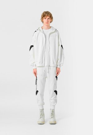 Sudadera blanca con capucha y bordado de alas negras, combinada con pantalones deportivos blancos con detalles a juego. Lleva botas de color claro.