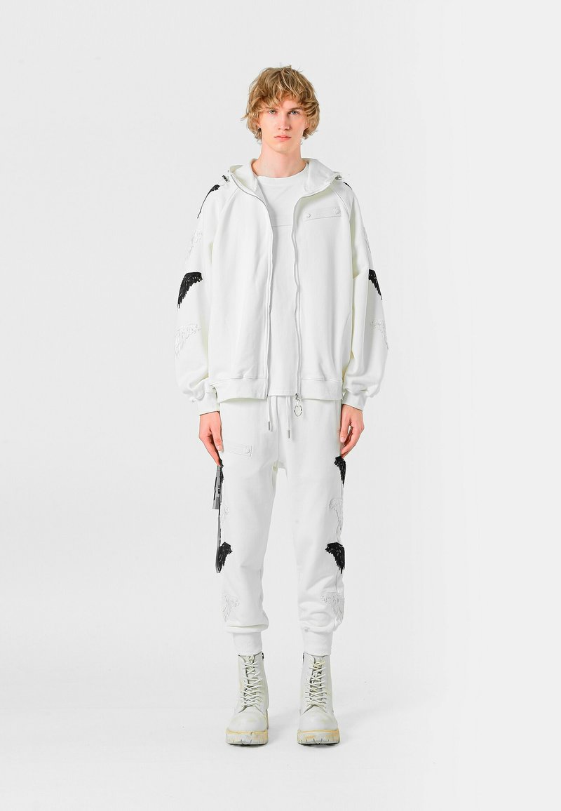 Sudadera blanca con capucha y bordado de alas negras, combinada con pantalones deportivos blancos con detalles a juego. Lleva botas de color claro.