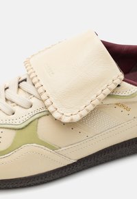 Baskets en cuir crème avec un rabat rembourré, des accents vert olive, des détails perforés et une semelle en caoutchouc noir texturé.
