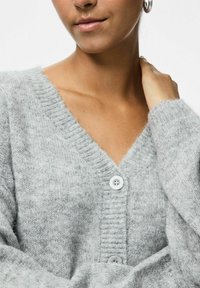 Sivi pleteni kardigan s V-izrezom, rebrastom teksturom i dva bijela dugmeta na prednjem dijelu. Mekani i lagani materijal.