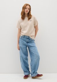 T-shirt en coton beige avec un petit graphique, associé à un jean en denim bleu clair ample et des baskets bordeaux avec une semelle en caoutchouc marron.