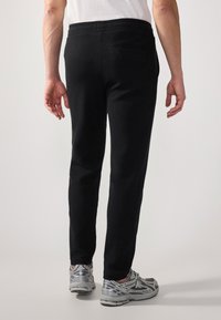 Schwarze, strukturierte Jogginghose mit enganliegendem Design und elastischem Bund. Verfügt über eine Gesäßtasche zur Aufbewahrung. Kombiniert mit grauen Sneakers.