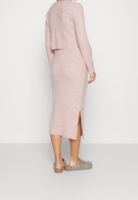 Kvinna iklädd en ljusrosa ribbad stickad långärmad topp och matchande midi kjol med ett sidoflik, kombinerat med beigea slip-on skor.