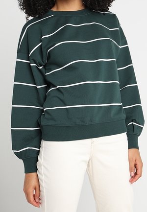 Femme portant un sweat-shirt vert foncé à rayures horizontales blanches et un pantalon blanc cassé, debout devant un fond clair uni.