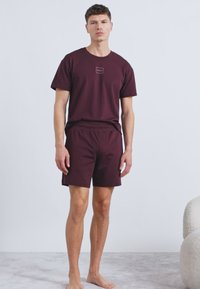 Burgundinpunainen lyhythihainen t-paita, jossa pieni logo rinnassa, yhdistetty samansävyisiin shortseihin. Kangas näyttää sileältä ja istuvuus on rento.