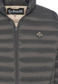 Grå dunjakke med vandrette syninger, en høj krave og en lynlås foran. Har et logopatch i diamantform på venstre bryst.