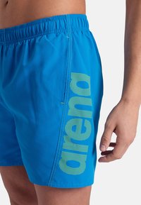 Pantaloni da nuoto blu con cintura elastica, tasche laterali, e un grande logo "arena" verde in un motivo a pois sul lato.