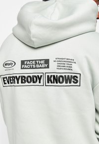 Sudadera verde claro con capucha ajustable y un distintivo texto gráfico en la parte posterior en negro, que muestra una mezcla de declaraciones y marcas.