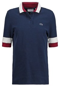Polo bleu marine avec col et poignets blancs et rouges, patte à deux boutons et petit logo crocodile sur la poitrine.